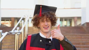 Red -haired curly guy with braces stands in a master's mantle and shows a thumb up. Curly graduate with braces smiles near the college video