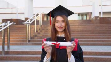 A university female graduate stands in a master's mantle and holds a diploma in front of her video