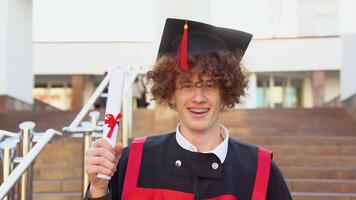 un tonto sonriente rojo -peludo Rizado chico con tirantes soportes en un maestría manto y sostiene un diploma. Rizado graduado con tirantes sonrisas cerca el Universidad video