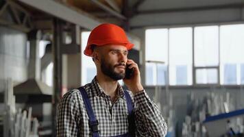 A young worker in a helmet at a large metalworking plant speaks on the phone. Professional worker in the finished product warehouse makes an order video