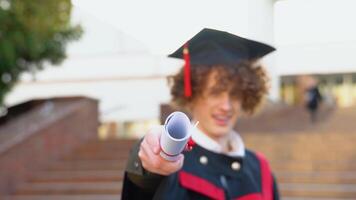 un joven rojo -peludo Rizado chico con tirantes soportes en un maestría manto participación un diploma estirado adelante a cámara. cámara enfocado en un diploma video