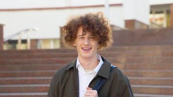 Outdoor portrait of cheerful red -haired curly student with backpack and books standing near college building, looking at camera and smiling video