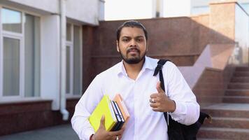 Happy Indian student stands on the stairs, showing a thumb up, in a university campus video