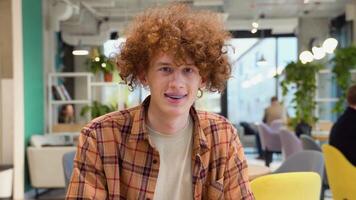 uma jovem vermelho -cabelo encaracolado homem com suspensórios senta dentro uma cafeteria sorrisos e parece às a Câmera video