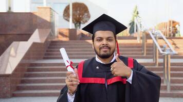 feliz indiano aluna graduado carrinhos em a escadas com diploma, mostrando uma polegar acima, dentro uma universidade campus video