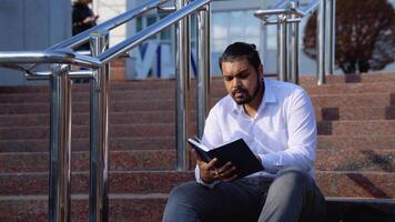 indio estudiante se sienta en el escalera lee libros en un Universidad instalaciones video