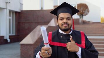 Happy indian student graduate stands on the stairs with diploma, showing a thumb up, in a university campus video