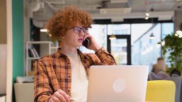 Young freelancer or blogger in glasses with braces sitting in a cafe talking on mobile phone while working on a laptop. Smiling programmer working on laptop sitting in a cafe video