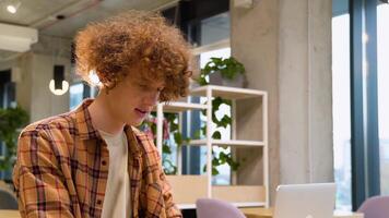 Young red -haired curly man sitting at a table in a cafe and working on a laptop, wears yellow shirt, creates new content for his blog, enjoys the work of a freelancer video