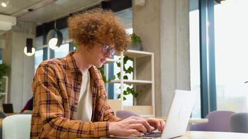 Young programmer sitting at a table in a cafe and working on a laptop, wears yellow shirt, creates new content for his blog, enjoys the work of a freelancer video