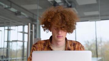 Funny redhead man straightens the hairstyle and using laptop sitting in office. Teenager freelancer distantly working or studying on computer typing online video