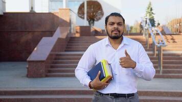 Happy Indian student woth books stands on the stairs, showing a thumb up, in a university campus video