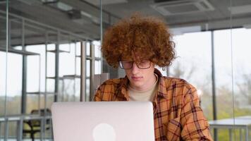 Young programmer sitting at a table in office and working on a laptop, wears yellow shirt, creates new content for his blog, enjoys the work of a freelancer video