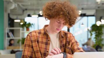 jovem vermelho -cabelo encaracolado homem sentado dentro uma cafeteria usa inteligente Assistir enquanto distante trabalhando video