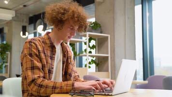 Tired disappointed programmer sitting at a table in a cafe and working on a laptop, wears yellow shirt. Young blogger creates new content for his blog, enjoys the work of a freelancer video