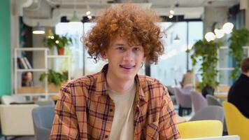 A young red -haired curly man with braces sits in a cafe smiles and looks at the camera video