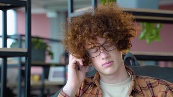 Young red -haired curly student freelancer listens to music in co working space while sitting on comfy soft armchair video