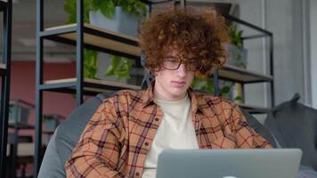 Young redhead blogger or freelancer with headphones holding notebook browsing chatting while sitting on comfy soft armchair wears yellow shirt in co working space video