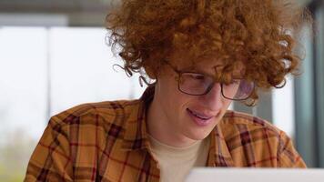 Focused redhead man in glasses using laptop sitting in office, serious teenager freelancer distantly working or studying on computer typing online video