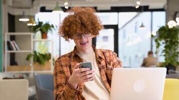 rojo -peludo Rizado persona de libre dedicación en lentes con tirantes hojeada teléfono inteligente y sonriente mientras sentado a mesa con ordenador portátil en ligero café en tiempo de día video