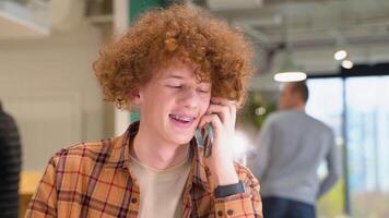 jong rood -harig gekruld Mens met een beugel zittend in een cafe pratend Aan mobiel telefoon terwijl ver weg werken video