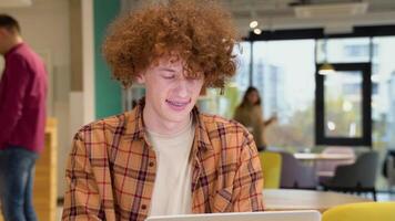 Young programmer with braces sitting at a table in a cafe and working on a laptop, wears yellow shirt, creates new content for his blog, enjoys the work of a freelancer video