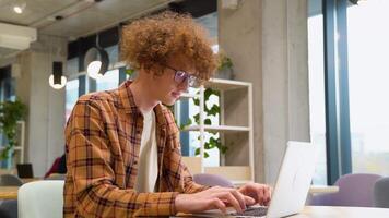 Tired disappointed programmer sitting at a table in a cafe and working on a laptop, wears yellow shirt. Young blogger creates new content for his blog, enjoys the work of a freelancer video