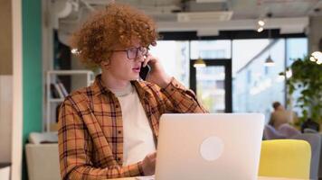 lächelnd Programmierer Arbeiten auf Laptop Sitzung im ein Cafe. jung Freiberufler oder Blogger im Brille mit Hosenträger Sitzung im ein Cafe reden auf Handy, Mobiltelefon Telefon während Arbeiten auf ein Laptop video