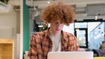 Tired freelancer or programmer during web working online. Red -haired curly hipster blogger sitting in cafe with laptop computer video