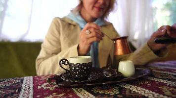kvinna häller turkiska kaffe från Cezve in i kopp. närbild långsam rörelse skott av kvinna hand med kopp på fyrkant tallrik, på tabell i Kafé utomhus. traditionell varm ofiltrerad kaffe eras i restaurang video