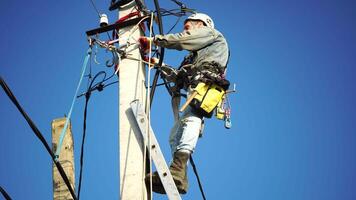 Skilled electrician in helmet fixes wires standing on ladder near high pole against blue sky on summer day backside view. Electrical service and mounting on the pole. Slow motion video