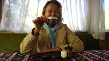 vrouw gieten Turks koffie van cezve in beker. detailopname langzaam beweging schot van vrouw hand- met kop Aan plein bord, Aan tafel in cafe buitenshuis. traditioneel heet ongefilterd koffie geserveerd in restaurant video