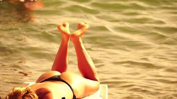 irreconhecível positivo mulher dentro bikini banhos de sol ou bronzeamento em beira-mar de praia durante verão. lento movimento do feliz turista desfrutando Sol bronzeado deitado em de praia cadeira salão às luxo recorrer video