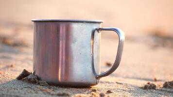 Close-up of a travel stainless mug with steaming hot tea standing on sandy beach at sunset. A concept of the world of beauty, nature and outdoor travel. Selective Focus. Slow motion video