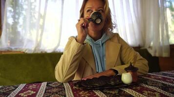 vrouw gieten Turks koffie van cezve in beker. detailopname langzaam beweging schot van vrouw hand- met kop Aan plein bord, Aan tafel in cafe buitenshuis. traditioneel heet ongefilterd koffie geserveerd in restaurant video