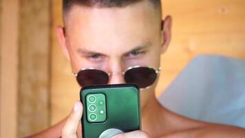 The young man in sunglasses, sunbathing by the sea and spending time on his mobile phone texting with girlfriend, looks at the camera in surprise, sees a news on the phone. video