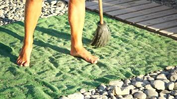 unrecognizable man sweeping the green carpet of artificial lawn on the beach from pebbles with broom. video