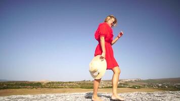 mujer en un rojo vestir y Paja sombrero en pie en un roca, disfrutando el Fresco aire. abrazando libertad y belleza de naturaleza. contento dama en parte superior de montaña con elevado manos mientras viento es soplo su vestido. video