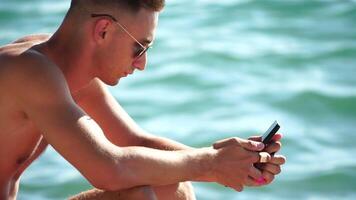 el joven hombre en Gafas de sol, broncearse por el mar y gasto hora en su móvil teléfono mensajes de texto con novia, mira a el cámara en sorpresa, ve un Noticias en el teléfono. video