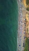 Aerial view of sandy beach, swimming people in sea bay with transparent blue water at sunset in summer. Happy People Crowd Relaxing On Beach. Holiday recreation ocean nature concept. Vertical video