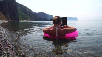 Woman freelancer works on laptop swimming in sea on pink inflatable ring. Happy tourist in sunglasses floating on inflatable donut and working on laptop computer in calm ocean. Remote working anywhere video