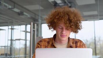 Funny redhead man straightens the hairstyle and using laptop sitting in office. Teenager freelancer distantly working or studying on computer typing online video
