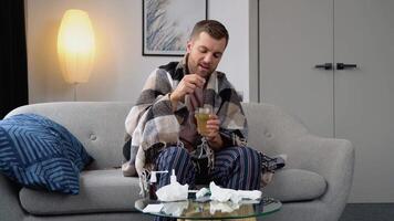 Health, flu and people concept - sick young man wrapped in a blanket drinking hot tea at home. man warming his hands. Heating season video