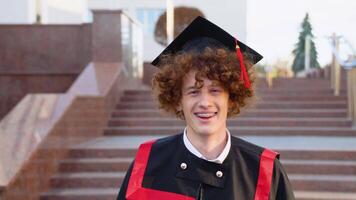 A young silly graduate in master's mantle. Curly graduate with braces smiles near the college video