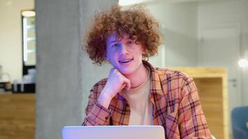 A young red -haired curly man with braces sits in a cafe smiles and looks at the camera. Young programmer in a cafe working on a laptop video