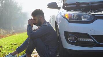 giovane uomo si siede vicino auto parlando su cellula Telefono perché il suo auto rotto fuori uso. caucasico maschio provando per chiamata amico quello essi Aiuto riparazione auto video