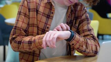 joven rojo -peludo Rizado hombre sentado en un café usos inteligente reloj mientras distante trabajando video