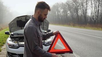 joven hombre preparando un rojo triángulo a advertir otro la carretera usuarios, coche Descompostura o motor fracaso detener en brumoso la carretera video