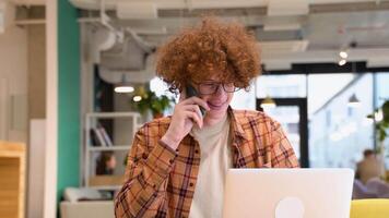 jung Freiberufler oder Blogger im Brille mit Hosenträger Sitzung im ein Cafe reden auf Handy, Mobiltelefon Telefon während Arbeiten auf ein Laptop. lächelnd Programmierer Arbeiten auf Laptop Sitzung im ein Cafe video