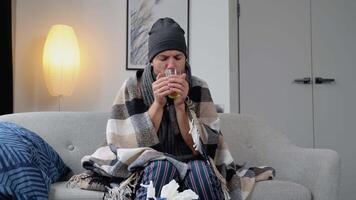 Health, flu and people concept - sick young man in hat wrapped in a blanket drinking hot tea at home. man warming his hands. Heating season video